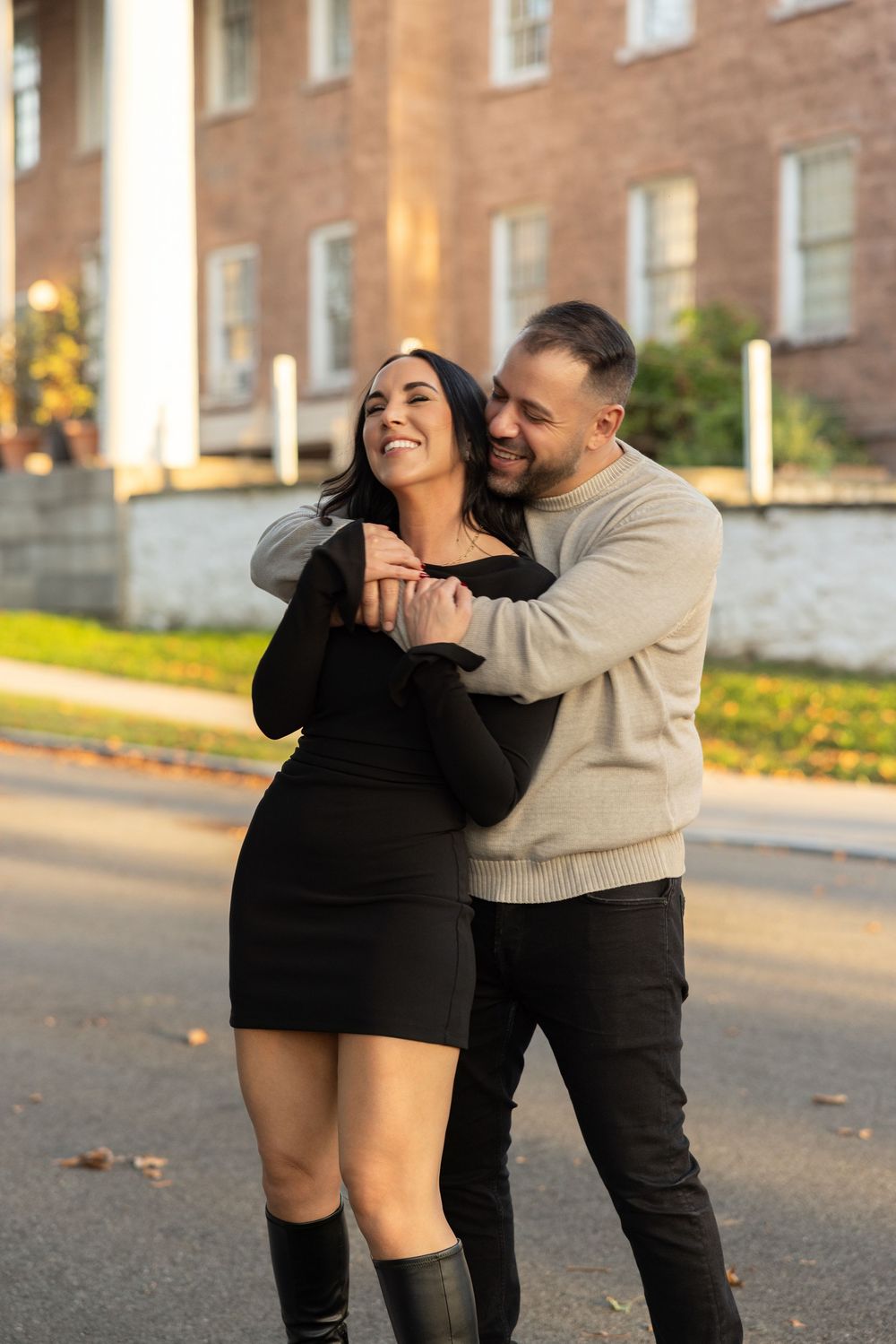A couple shares a tender embrace on a sunny afternoon in front of a brick building.