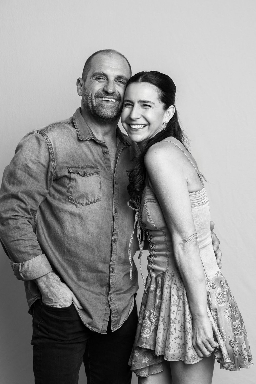 Black and white photo of a smiling couple posing together, man in denim shirt and woman in floral dress.