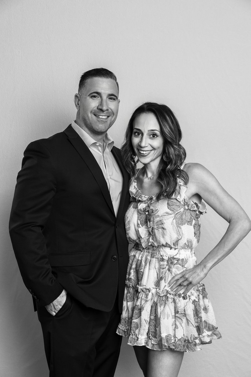 Black and white portrait of a smiling couple, man in suit and woman in floral dress, posing together.