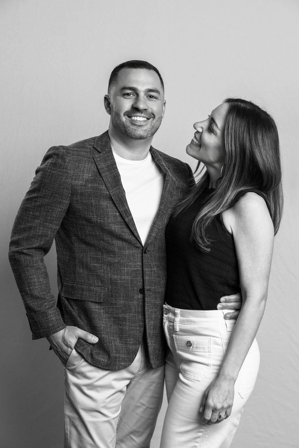 Black and white portrait of a smiling couple posing together, man in blazer, woman in black top and white jeans.