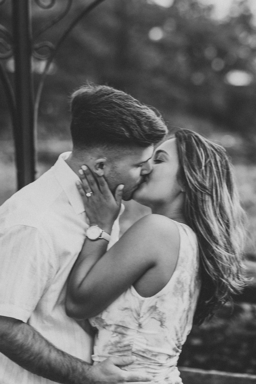 Black and white photo of a romantic couple sharing a passionate kiss outdoors, woman wearing a watch and ring.