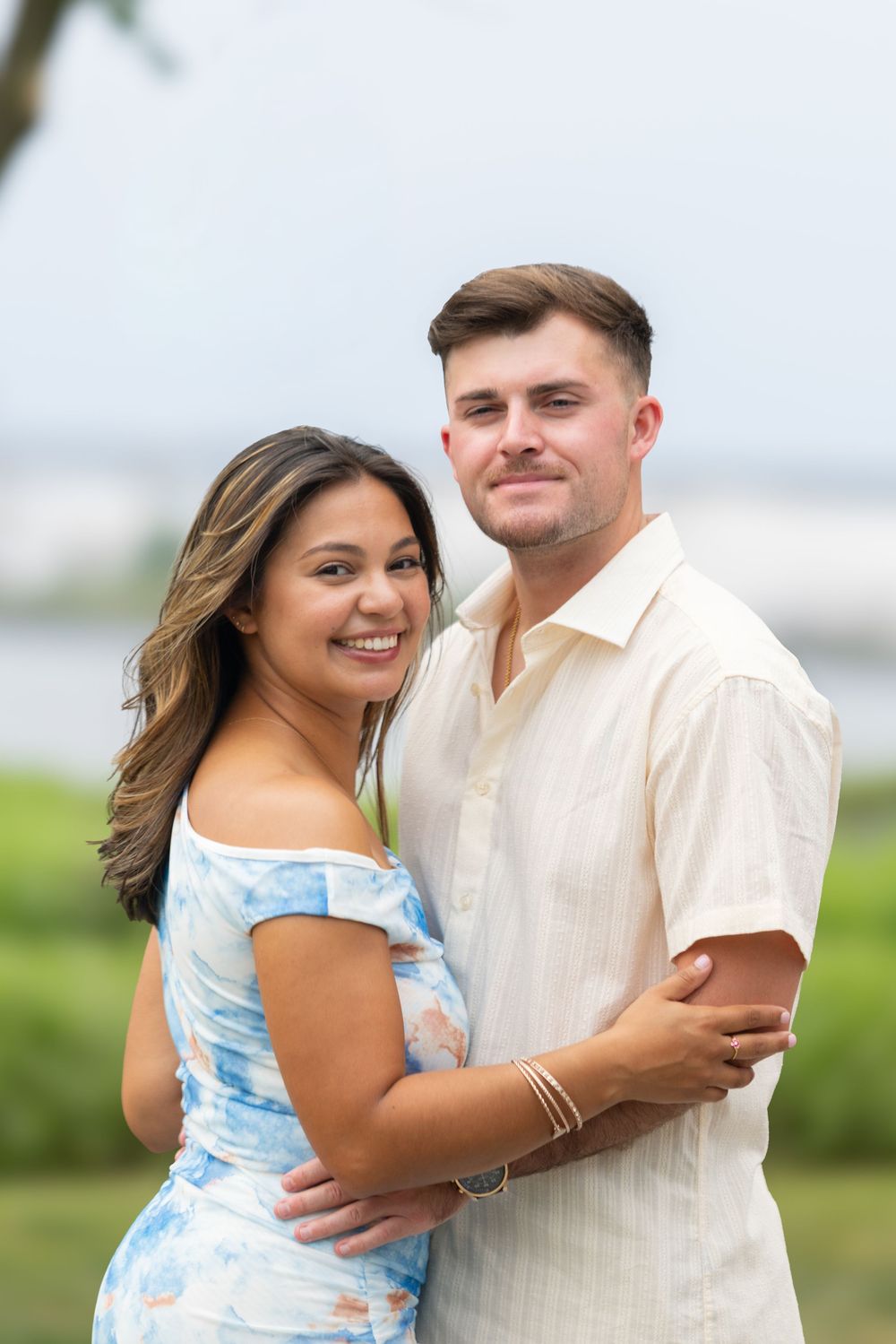 A smiling couple embraces outdoors, woman in floral off-shoulder dress, man in cream shirt, with green landscape behind.