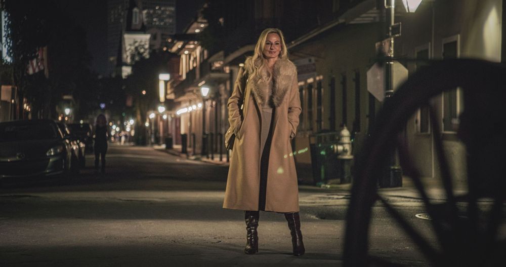 A person in a beige coat walks along a dimly lit street at night with buildings visible in the background.