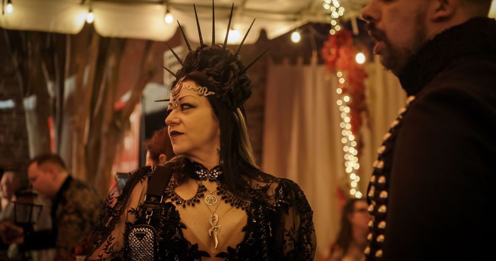 People in dark masquerade masks and formal attire gather at an elegant party with warm lighting and decorative elements.
