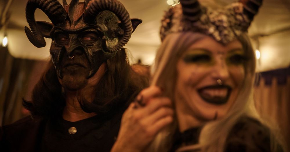 A group wearing ornate Venetian-style masks poses at a dimly lit formal event with string lights in the background.