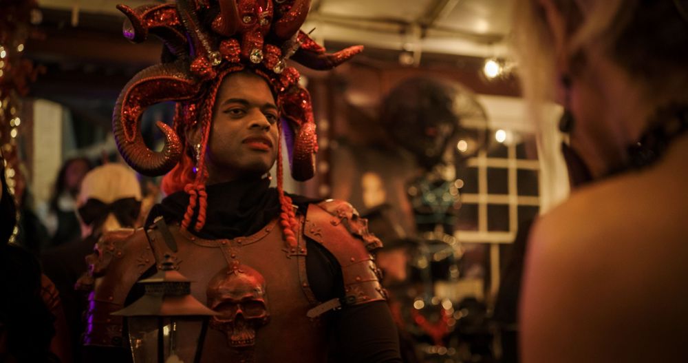 A performer wearing an elaborate red headdress and costume appears in a dimly lit indoor setting.