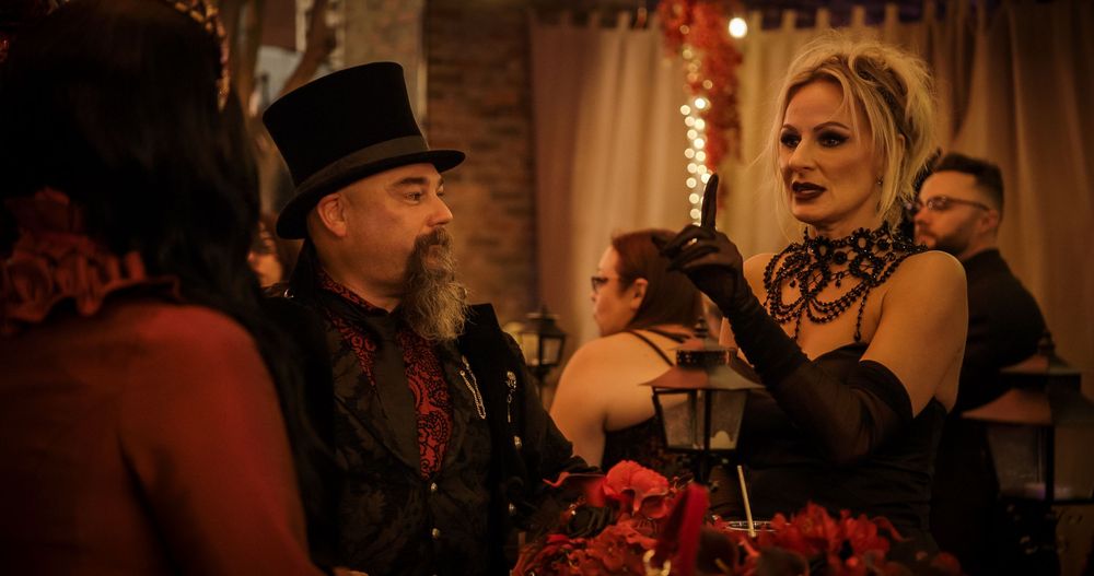 Two people in Victorian-style gothic attire converse at an elegant candlelit event with red decor.