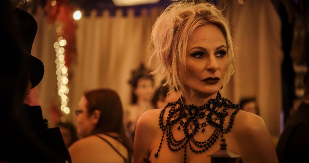 A person in a dark lace mask poses dramatically in warm golden lighting at an elegant evening event.