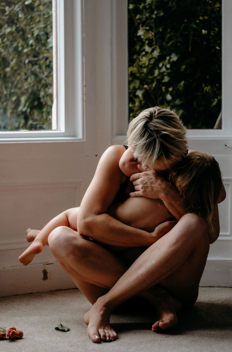 Mother sitting by a window cuddling her young child during an intimate natural motherhood photography session at home.