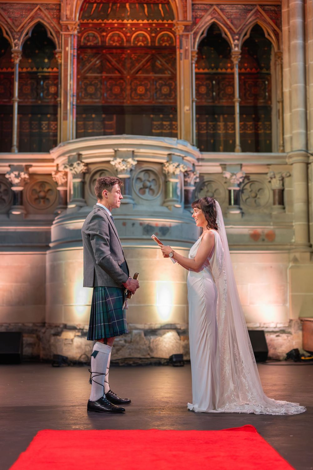 A couple in wedding attire share a romantic dance on a red carpet in an elegant venue with ornate architecture. Cottiers