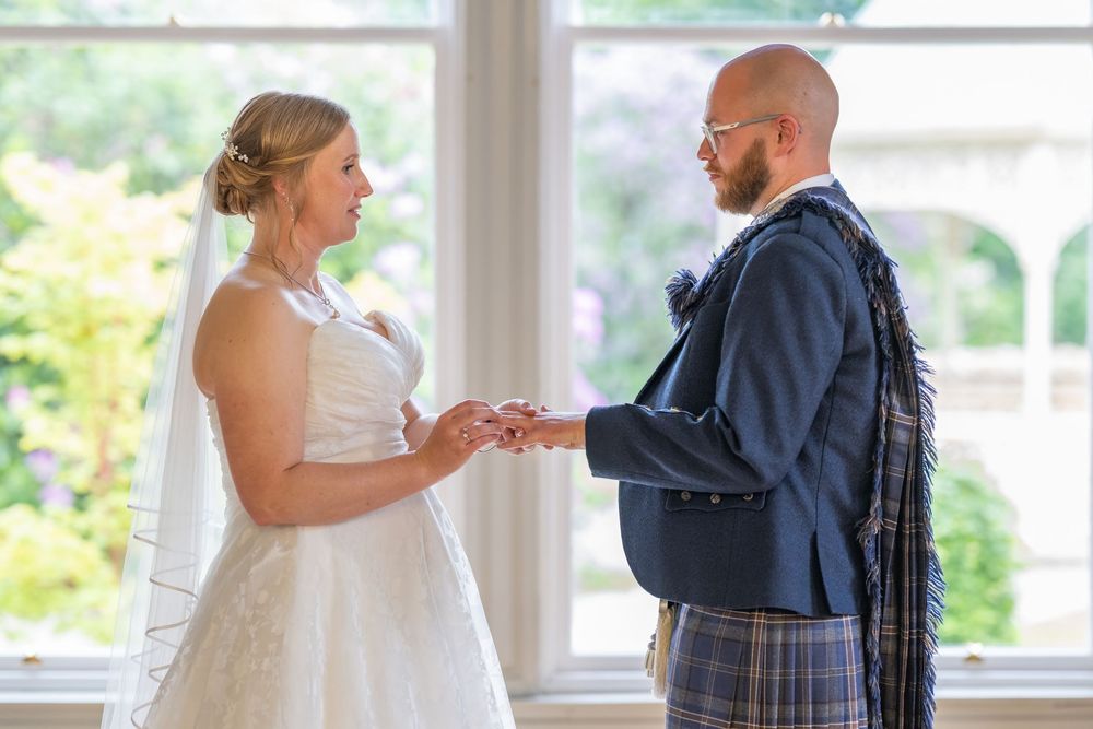 A couple in wedding attire share a tender dance moment in a bright sunlit room with windows.