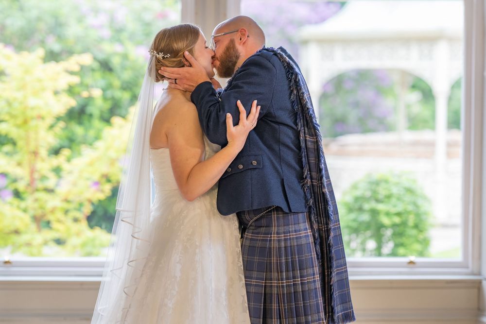 A couple shares a romantic kiss in front of large windows during their Scottish wedding ceremony.