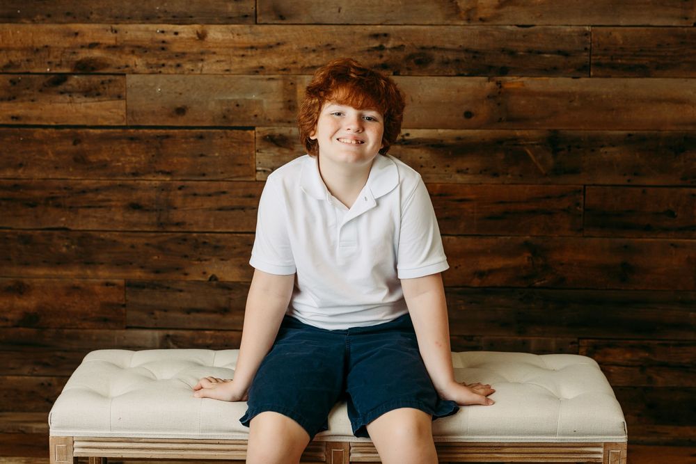 Indoor portrait against rustic wood paneled wall wearing casual white shirt and blue shorts.
