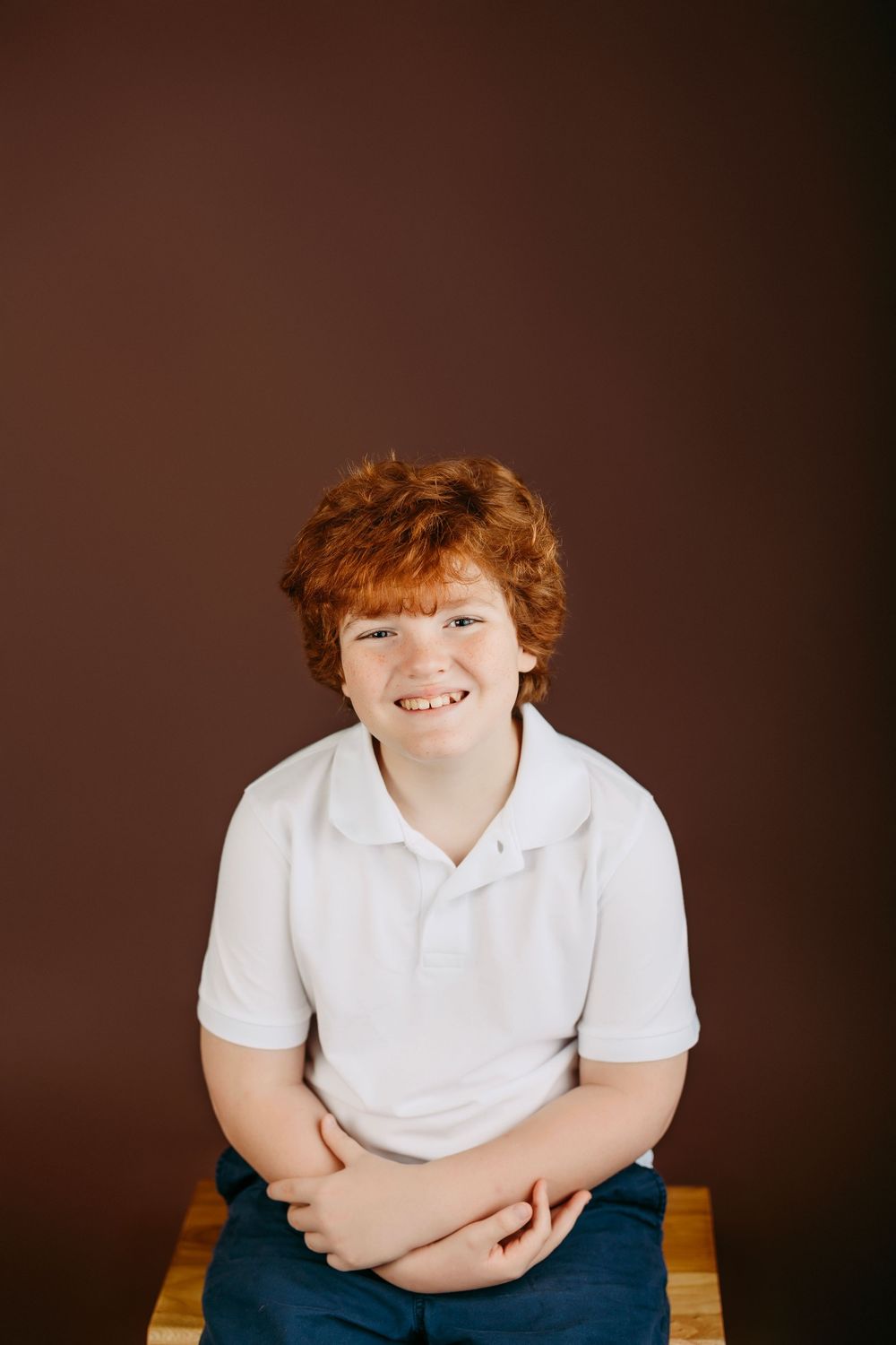 Studio portrait of a young person in a white polo shirt sitting on a wooden bench against a brown background.