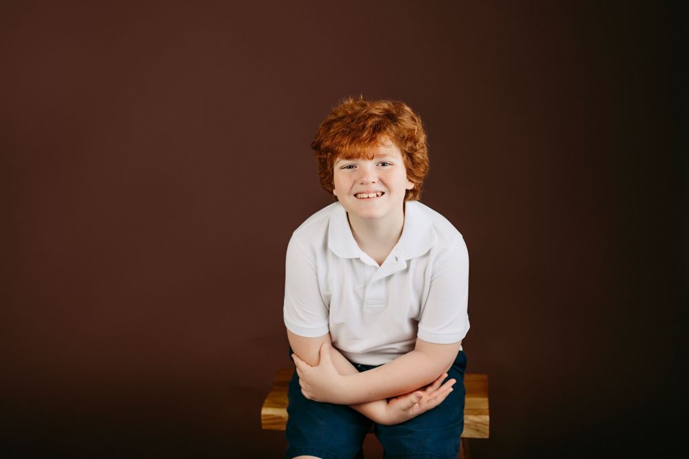Portrait of a smiling youth in white polo shirt seated on wooden bench with dark backdrop.