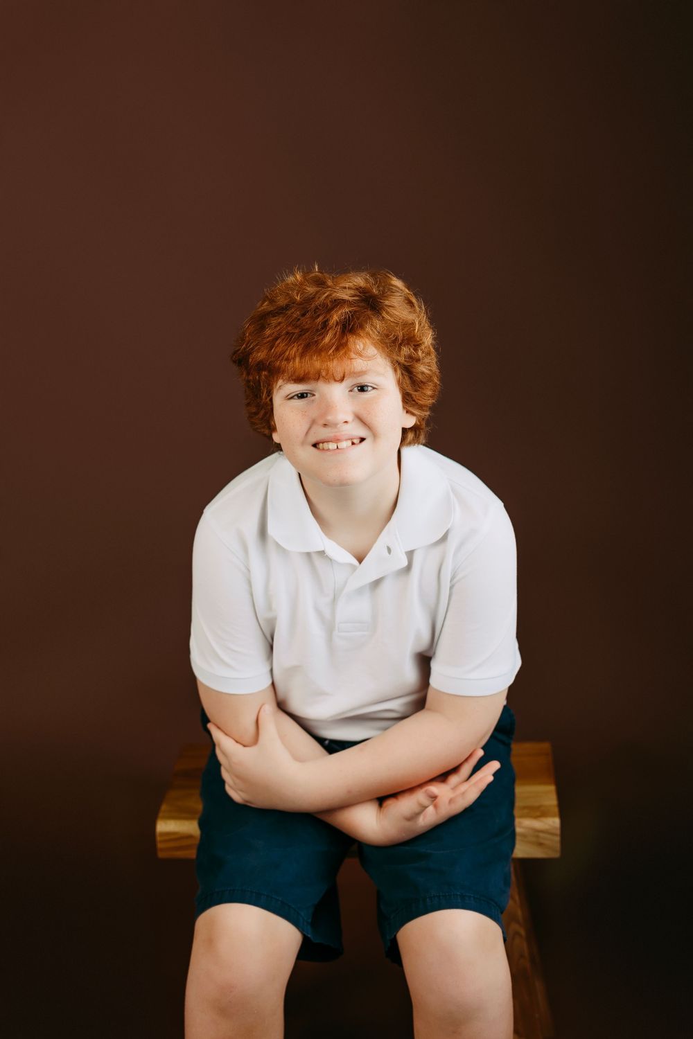Studio portrait of a child in a white polo shirt and dark shorts sitting on a wooden bench against a brown background.
