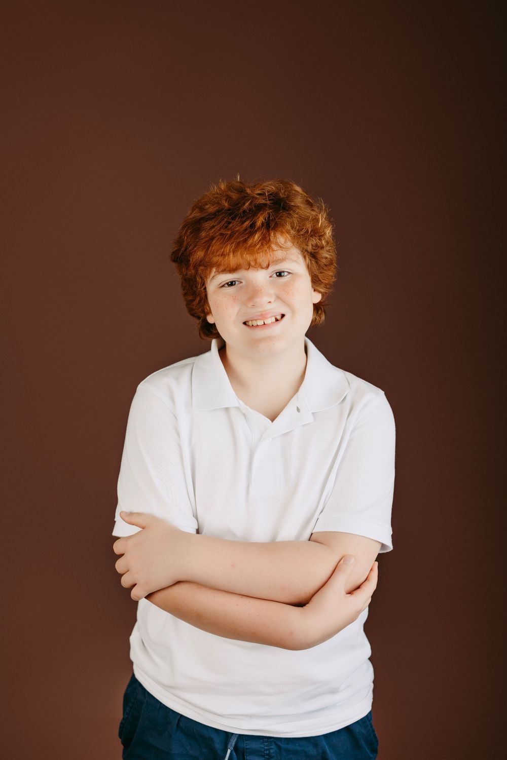 Studio photo of a person in white polo shirt with arms crossed against brown background.