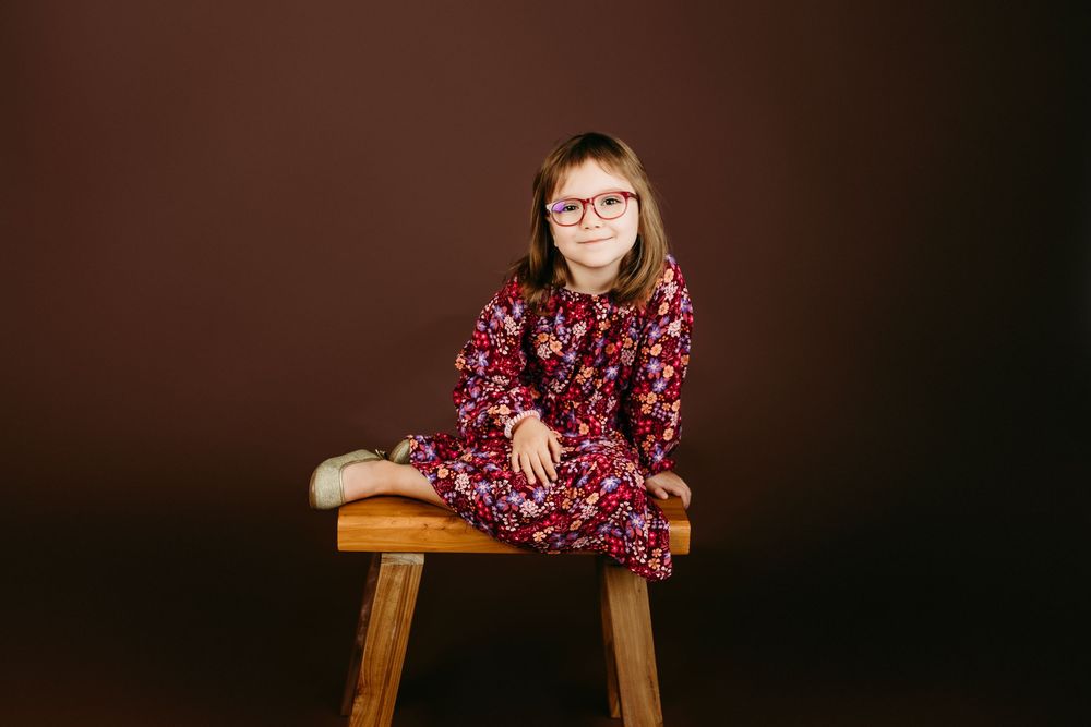 A child in a floral dress poses against a dark background while sitting on a wooden stool.