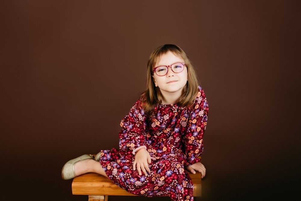 Young person in floral dress and glasses sitting on wooden bench against dark background.