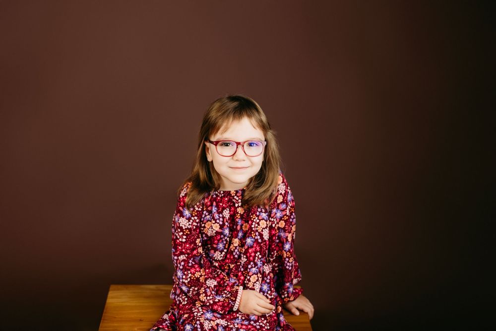 Portrait of someone wearing red floral dress and glasses sitting on wooden furniture.