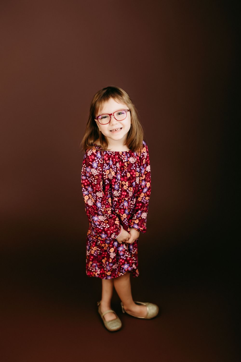 Young child wearing red floral dress poses on dark background.