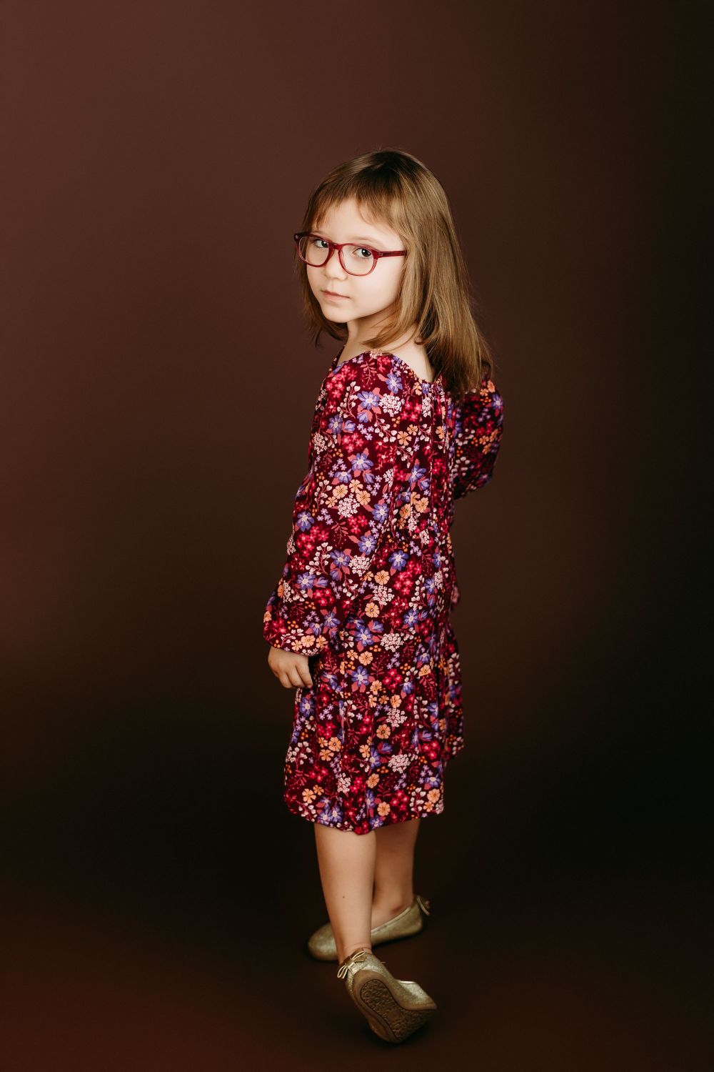 A child wearing a floral burgundy dress and glasses poses against a dark backdrop.