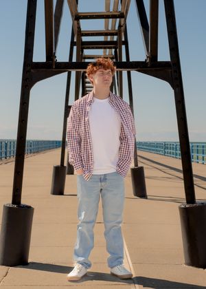 Person in checkered shirt and jeans standing under a pier structure on a beach.