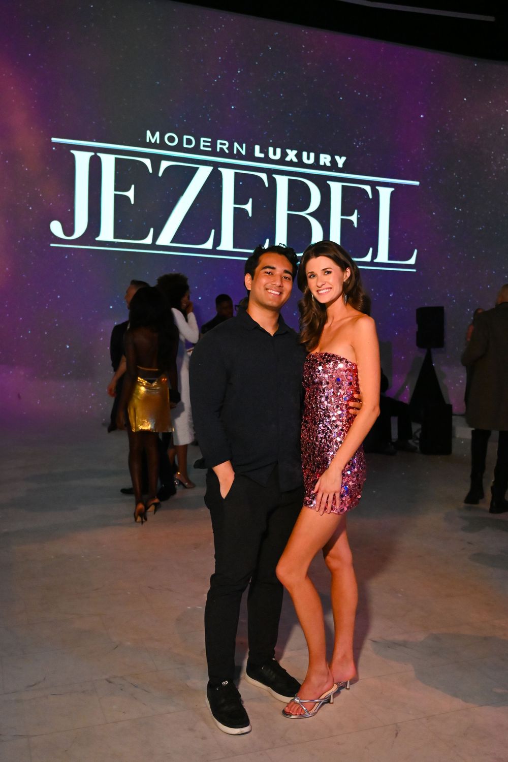 Two people pose together at a modern luxury fashion event with Jezebel branding visible in the background.