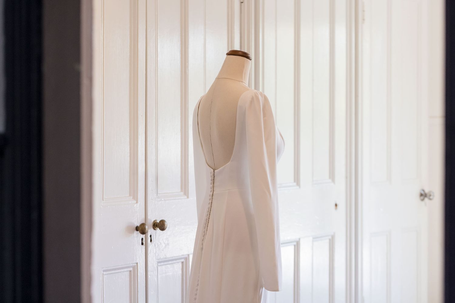 A simple white wedding dress with long sleeves hangs elegantly against white doors in natural light.