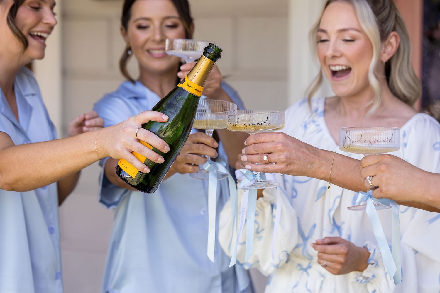 A Bride with her Bridesmaids in blue and white outfits stands together on a porch with a pink door and garden setting pouring champagne.