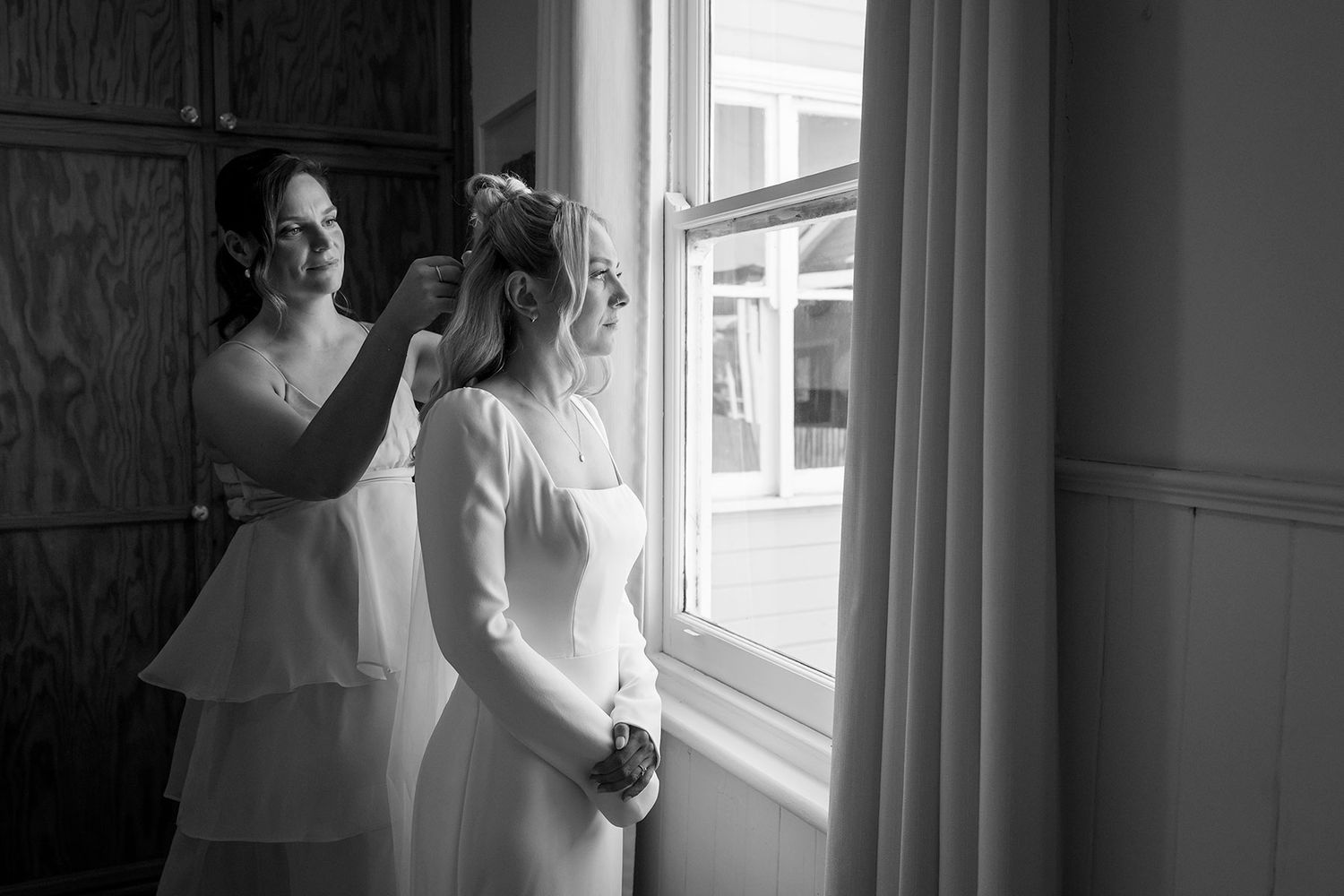 A bride and bridesmaid share an intimate moment by a window in black and white, helping the bride with her hair during preparations.