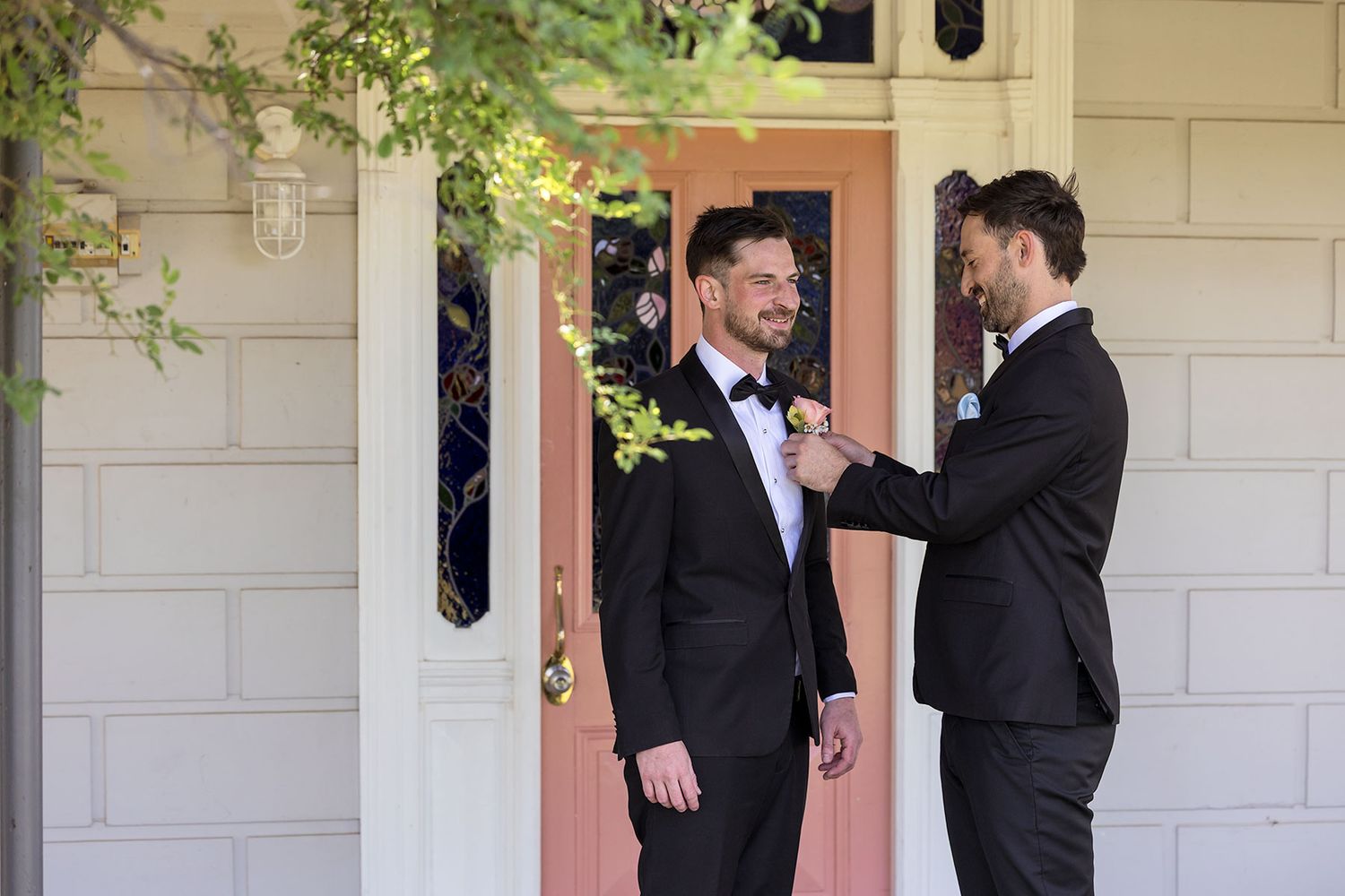A groomsman helps the groom with the button hole flower on his suit jacket on an old veranda with a pink door during wedding preparations.