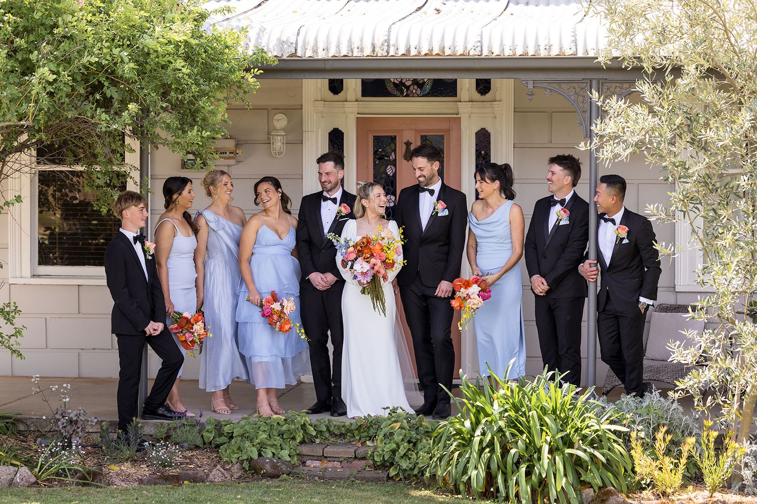 Wedding party in formal attire poses together outside in garden setting with bridesmaids in light blue dresses.