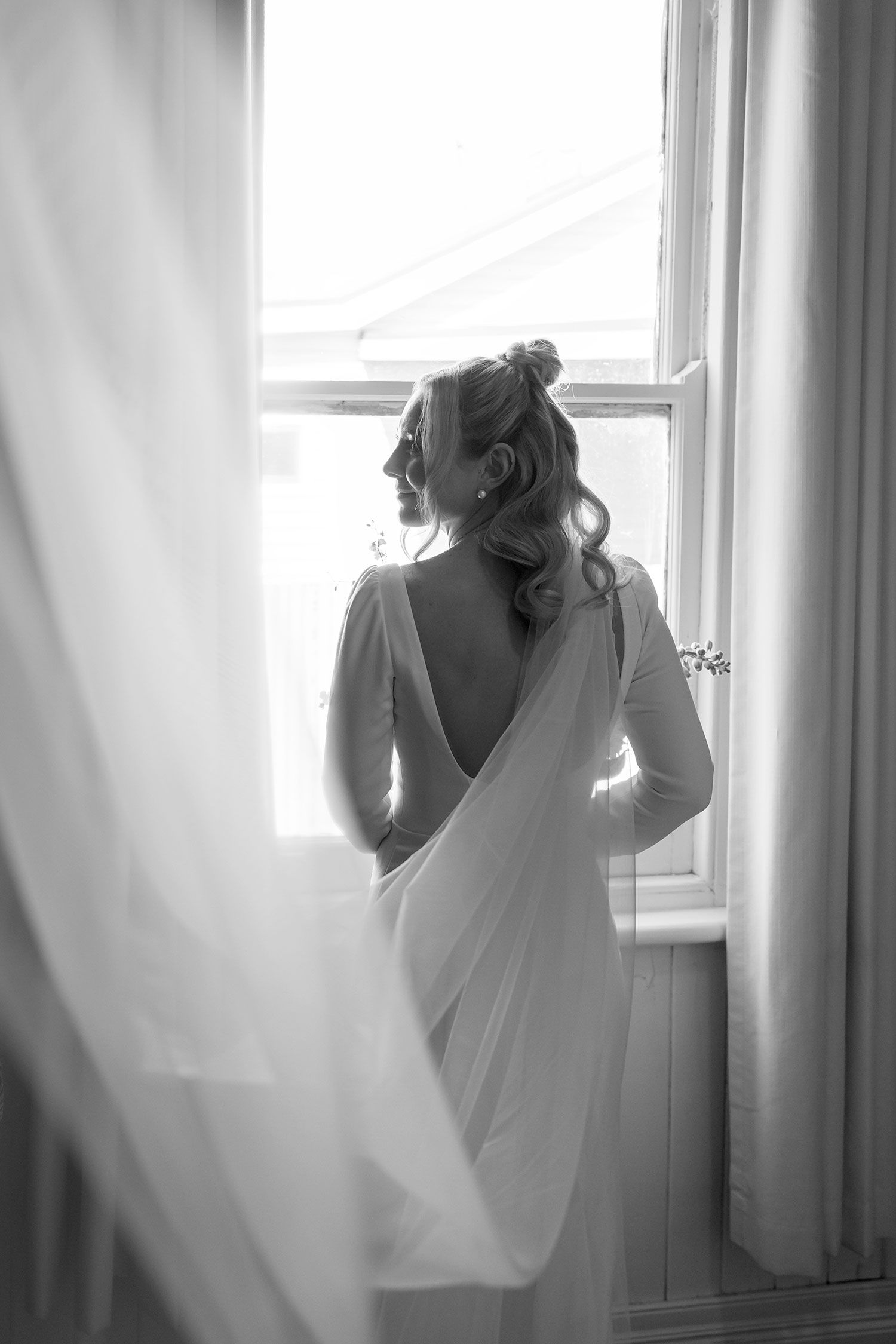 A bride in a flowing white dress stands silhouetted against a bright window in an elegant black and white photograph.