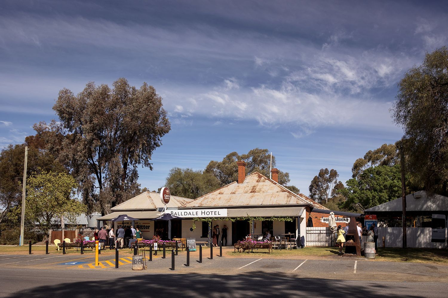 A panoramic view of a rural Australian outback Pub, the Balldale Hotel with parked vehicles and gum trees under a blue sky.