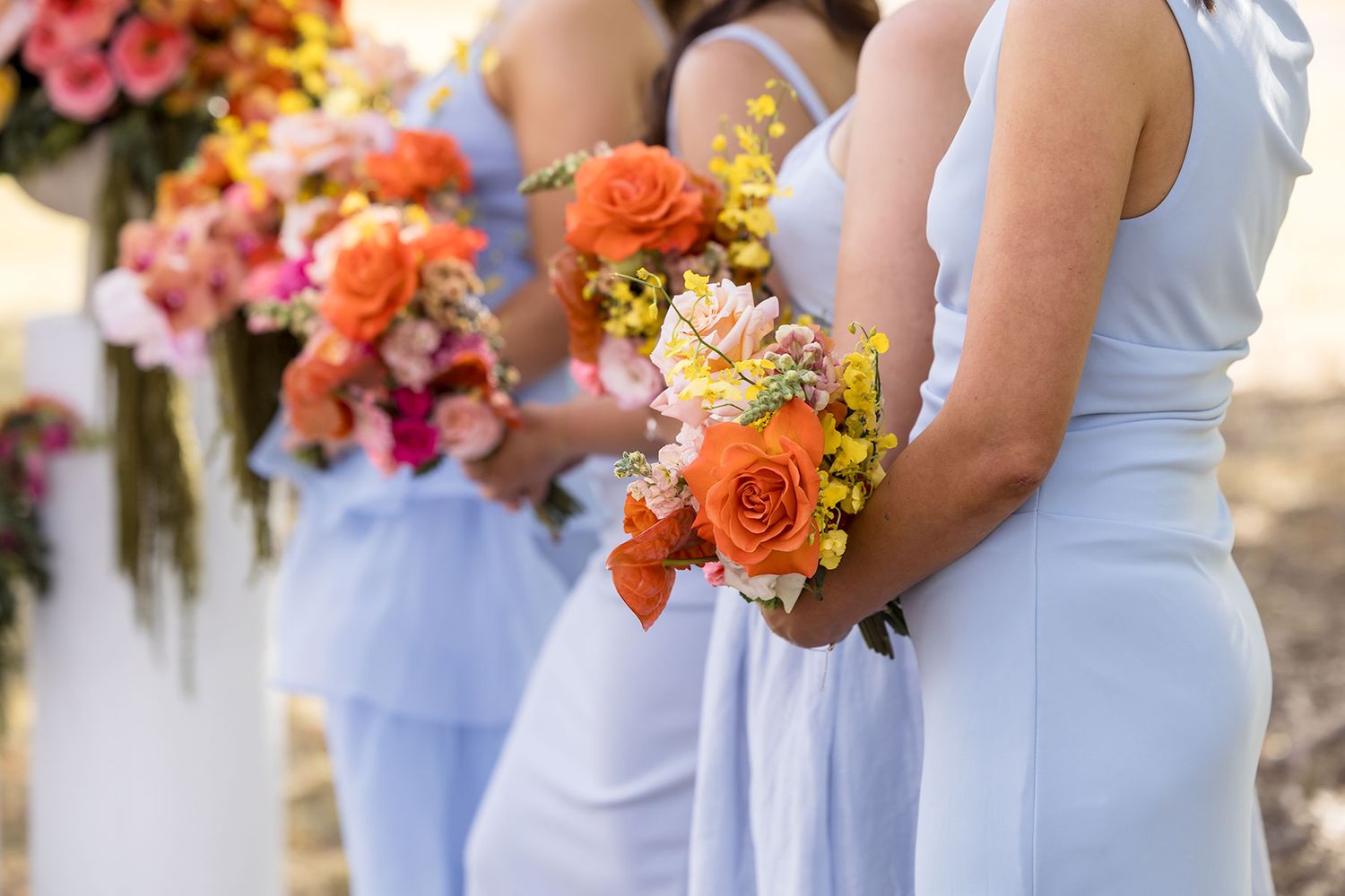 Bridesmaids in light blue dresses hold vibrant orange and coral floral bouquets during an outdoor wedding ceremony at the Balldale Hotel.