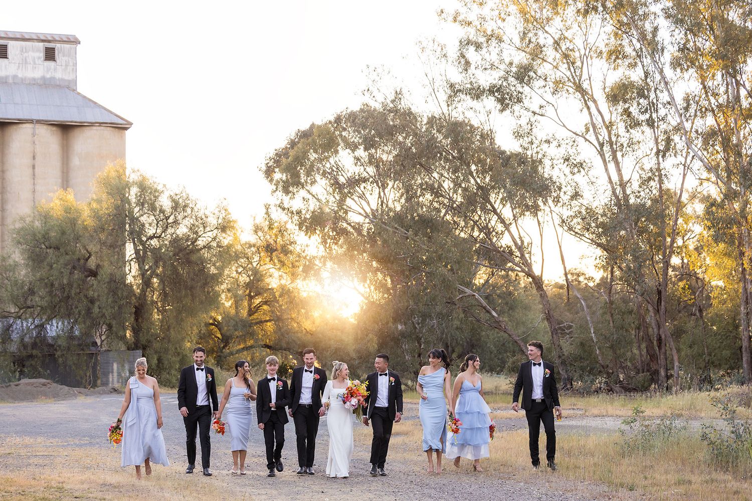 A Bride and Groom walking holding hands with their Bridal Party, with long grass and an old silo with the sun setting in the background.