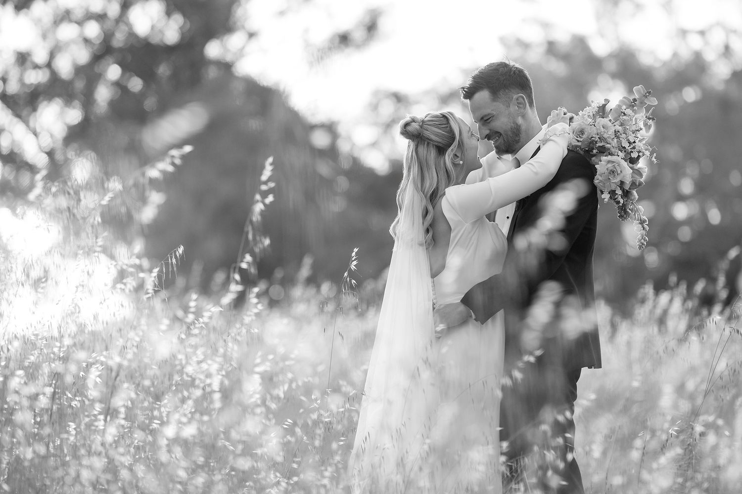 A black and white image of a couple sharing a romantic embrace in a sunlit meadow during their wedding photoshoot.