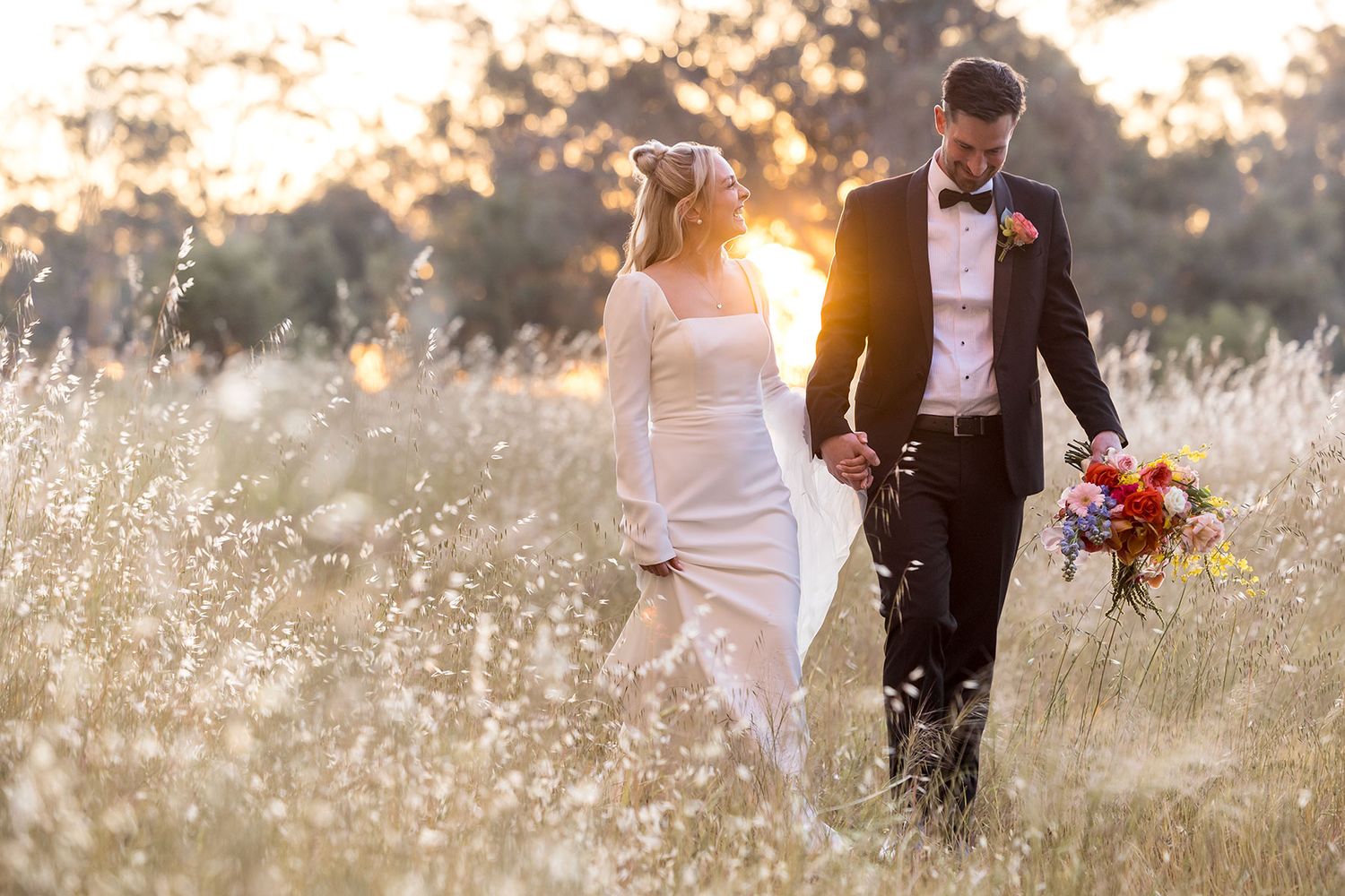 An image of a Bride and Groom walking in long grass holding hands with the sun setting in the background