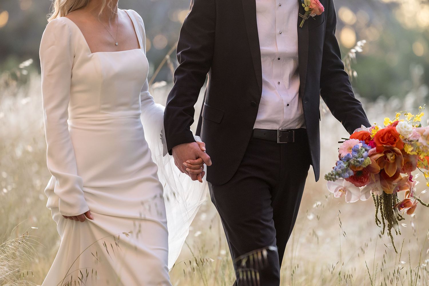 A close up image of a Bride and Groom walking in long grass holding hands with the floral bouquet and the sun setting in the background.