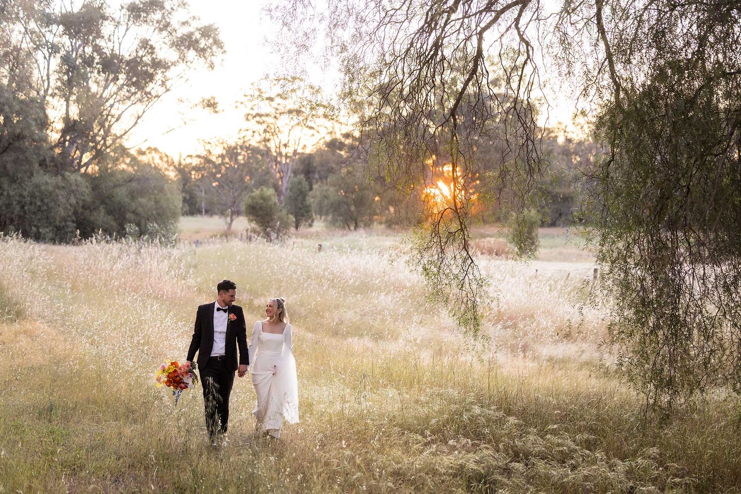 An image of a Bride and Groom walking in long grass holding hands with the sun setting in the background