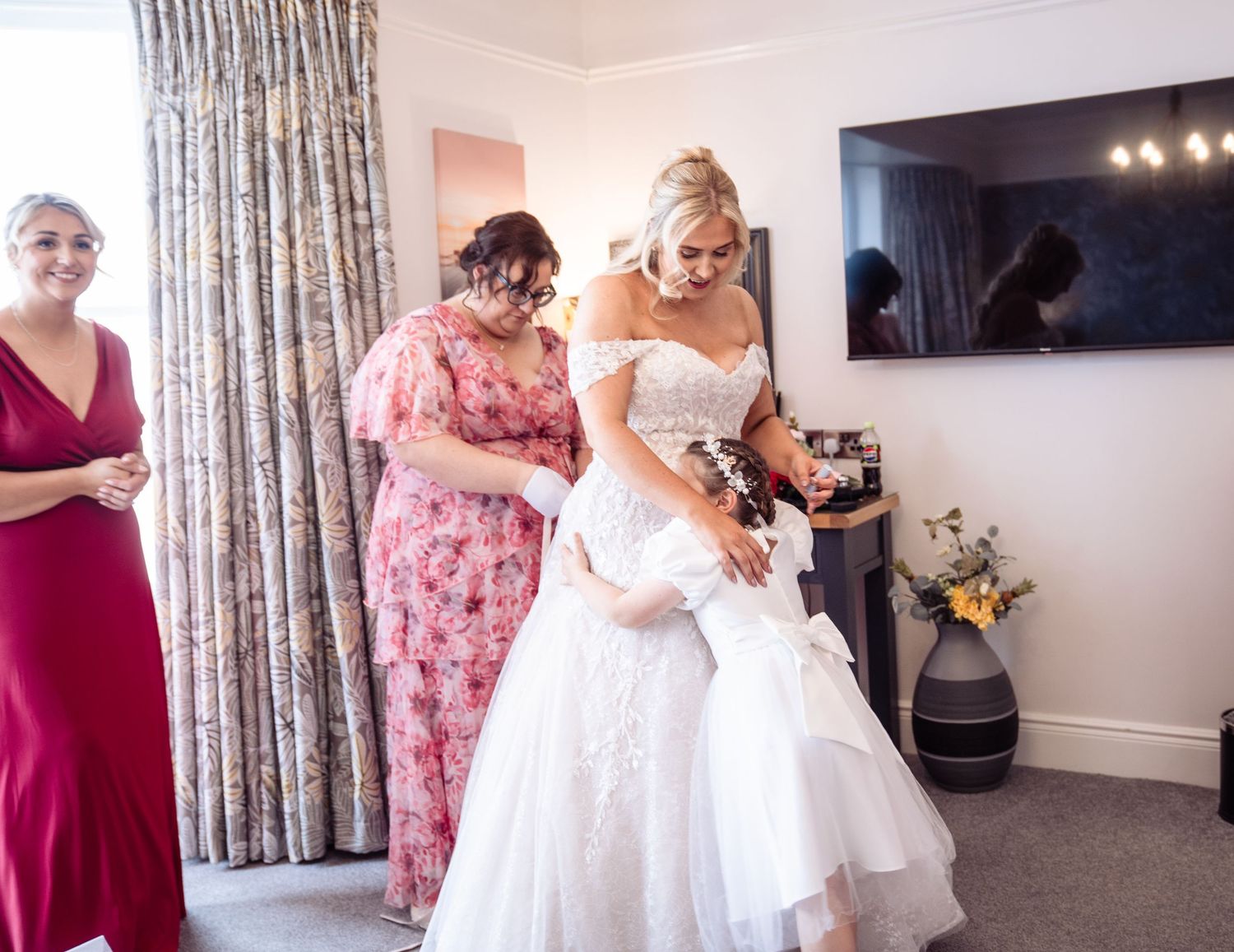 A bride in a white wedding dress gets ready with help from attendants in a room with floral curtains and a mounted TV.