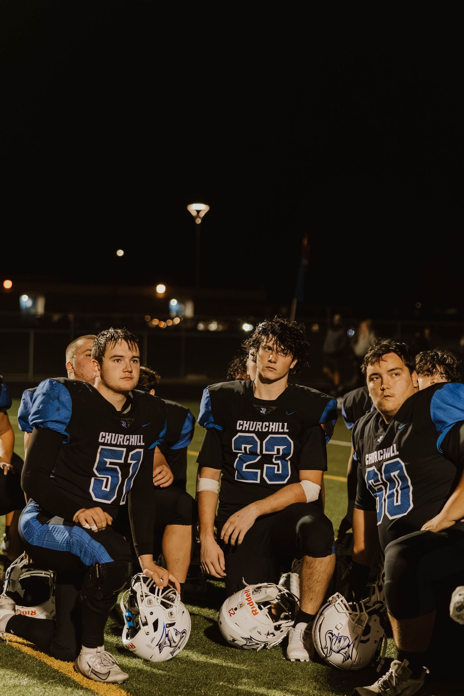 High school football players in blue and black uniforms sit together on the sidelines at night.