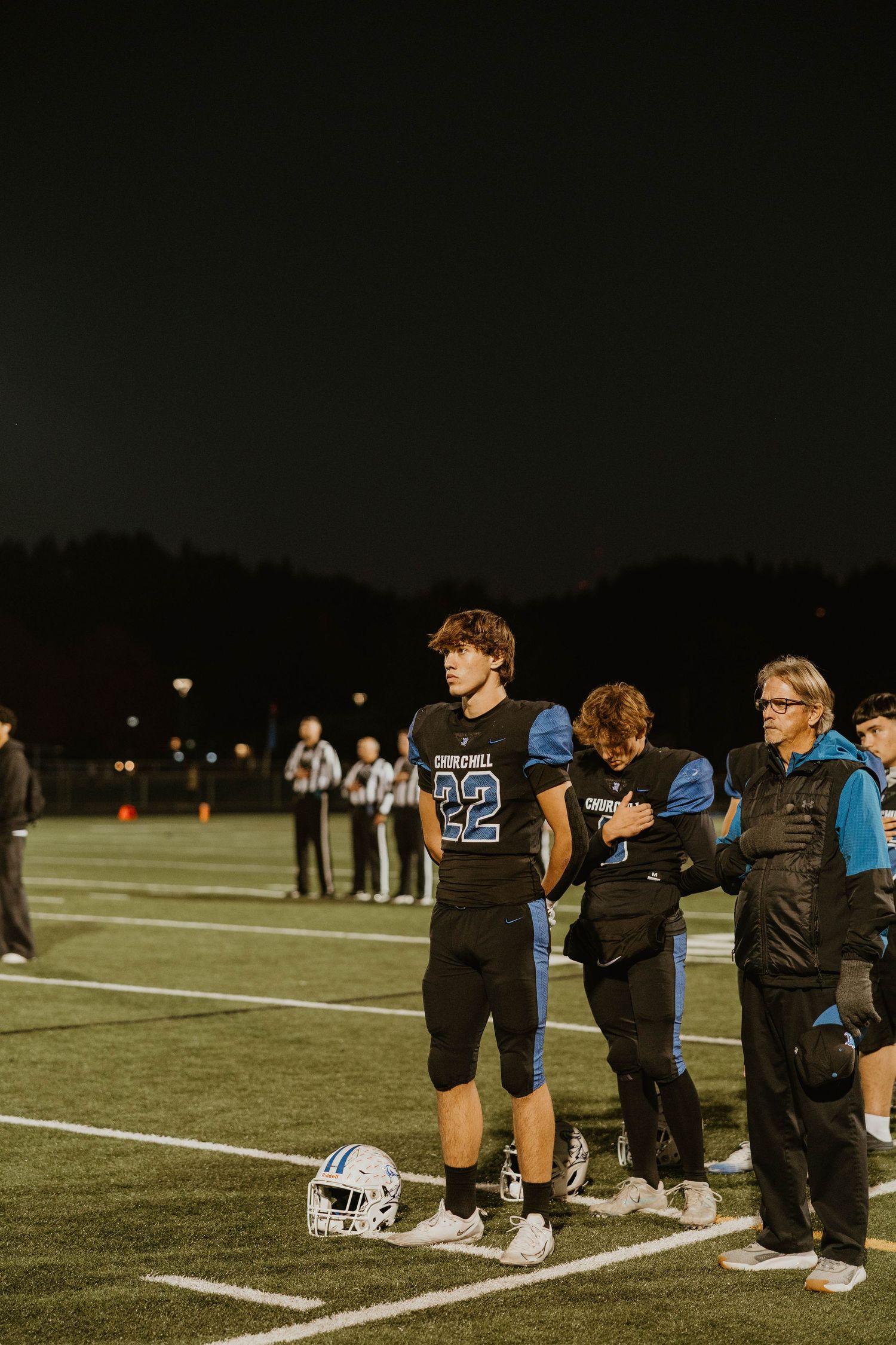 Football players kneel on the sideline during a nighttime game at a sports field.