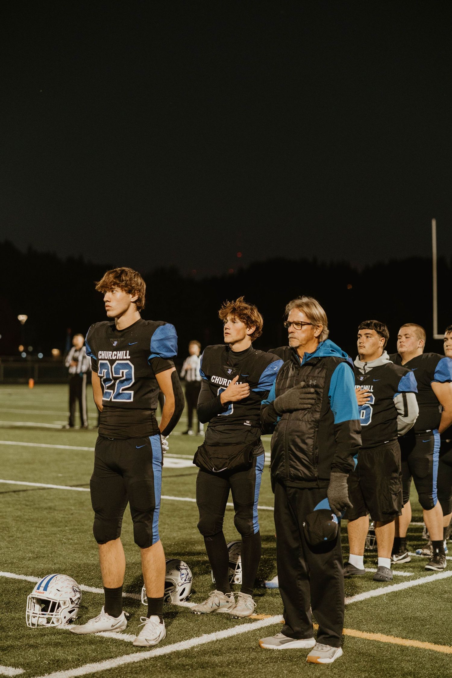 Youth football players kneel along the sideline during a night game under stadium lights.