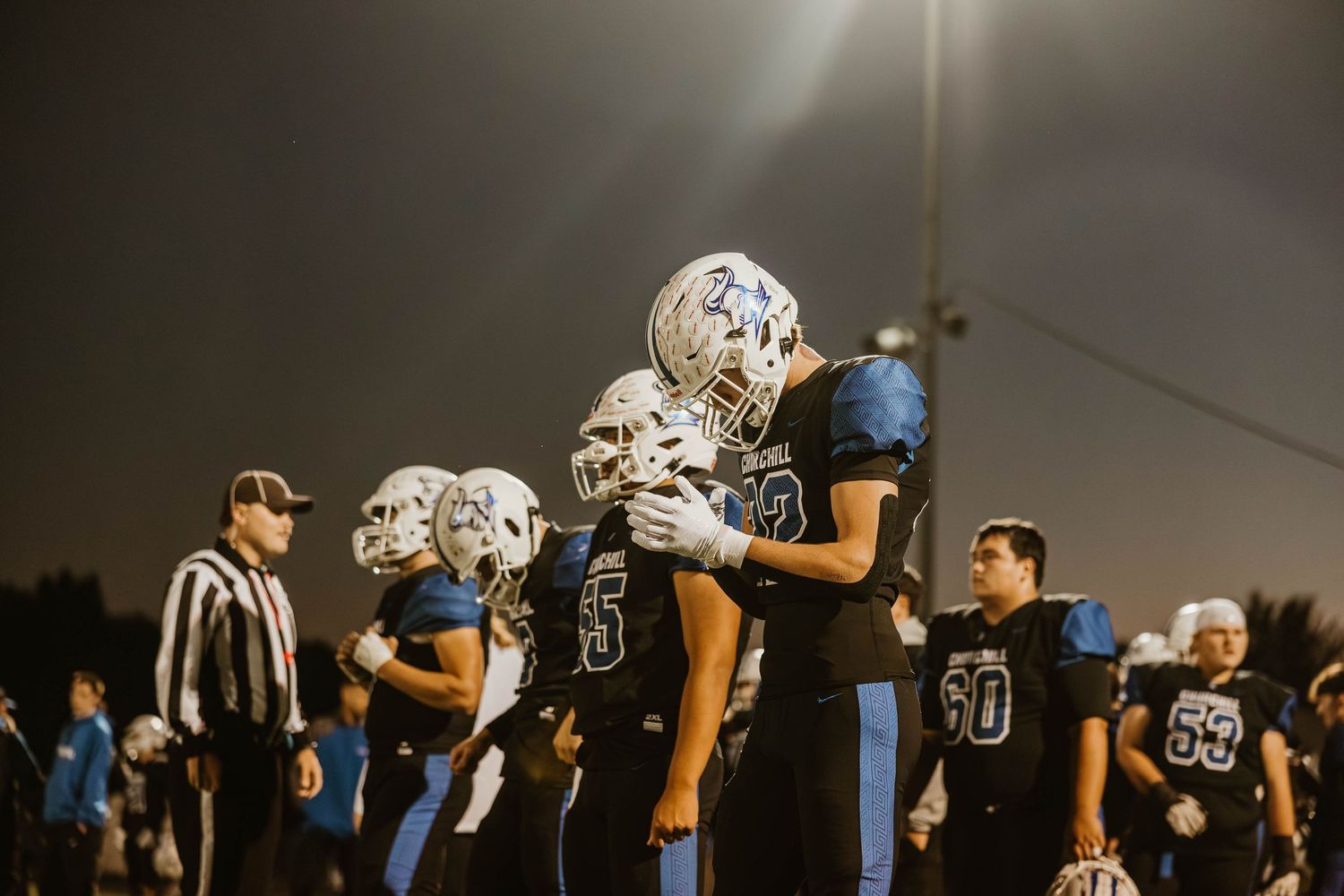 Football players in black and blue uniforms gather on the sidelines during a night game.