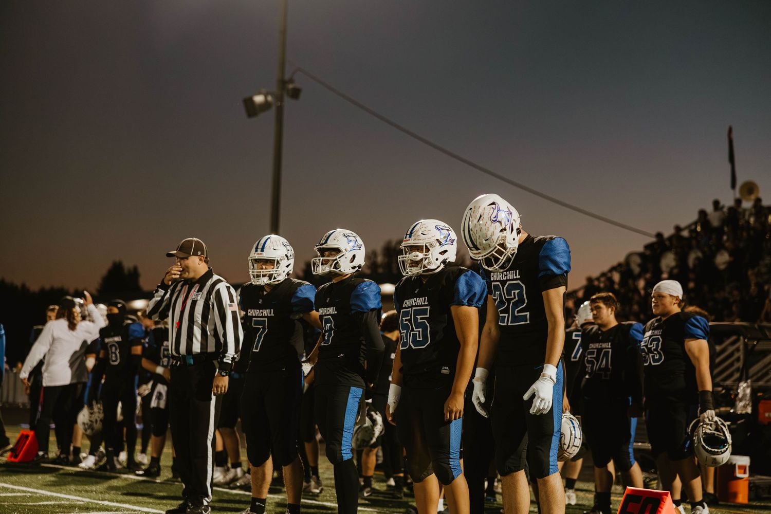 Football players and officials gather on the sideline during a night game under stadium lights.