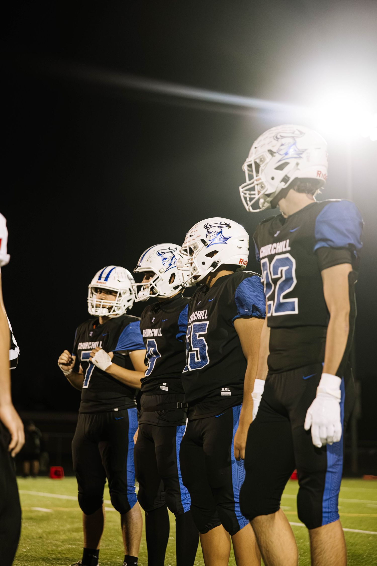 Football players in black and blue uniforms line up on the field during a nighttime game.