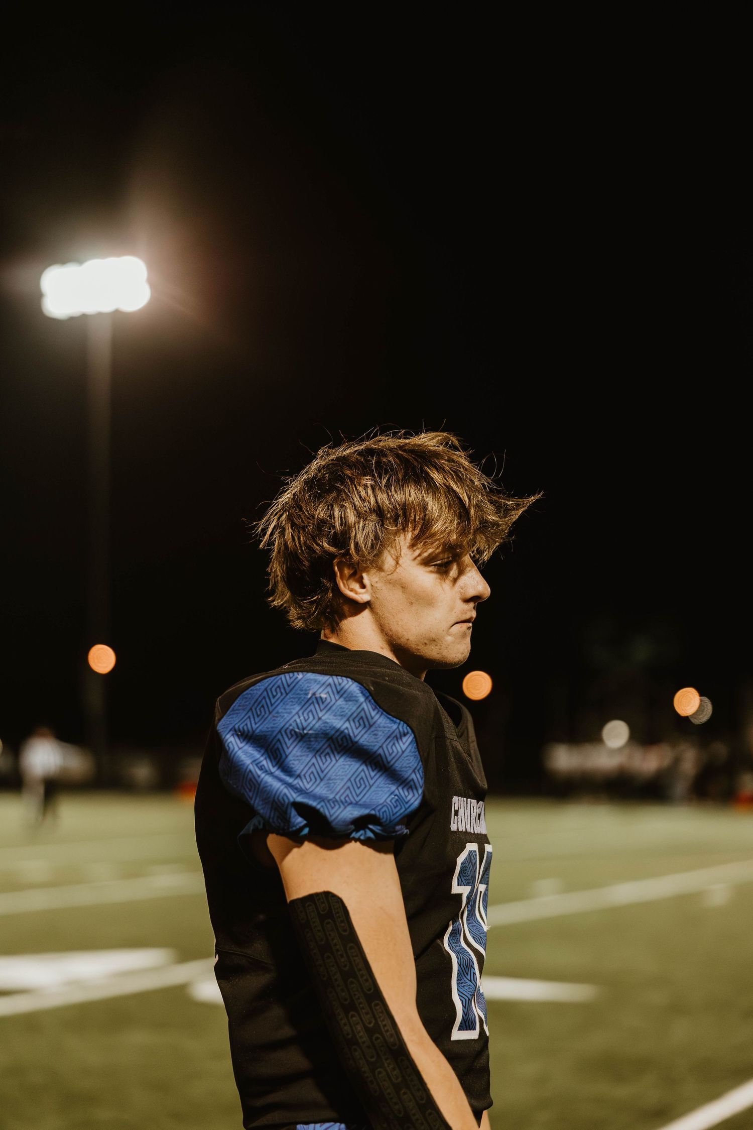 Football player in blue and black uniform stands under stadium lights during nighttime game.