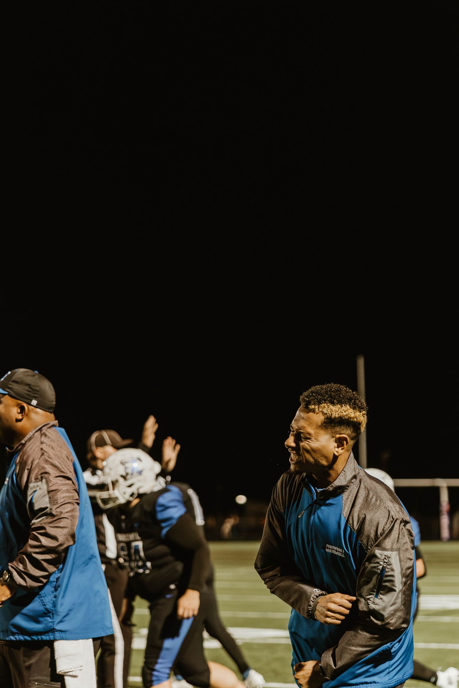 Coaches and staff members in blue jackets interact on the sidelines during a nighttime football game.